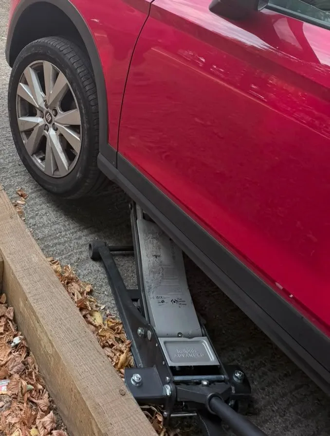 A red hatchback raised on a trolley jack beside a kerb, front wheel still on, ready for the tyre to come off